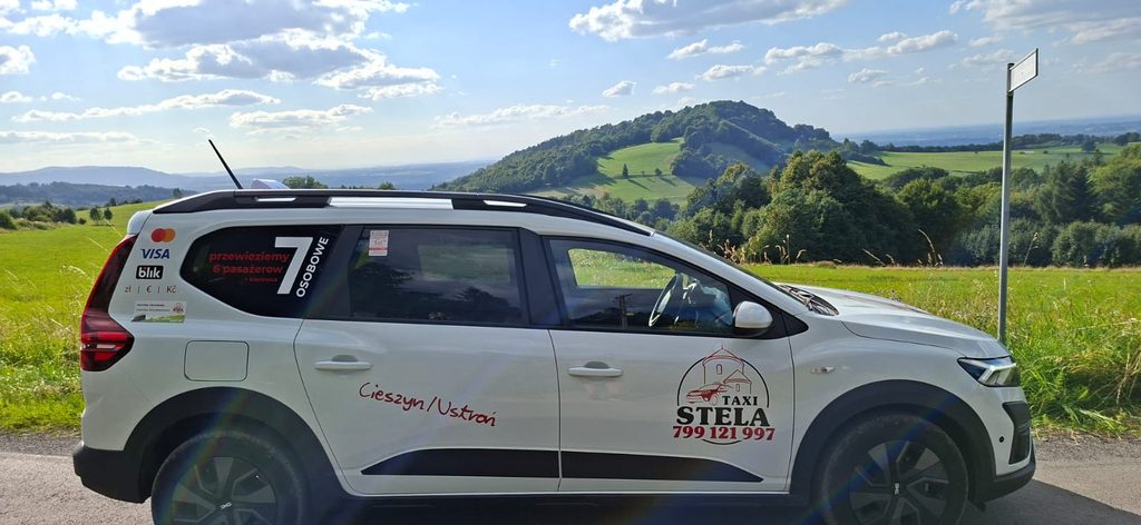 Biała Dacia Jogger Taxi Stela na tle zielonych wzgórz, pól i drzew.